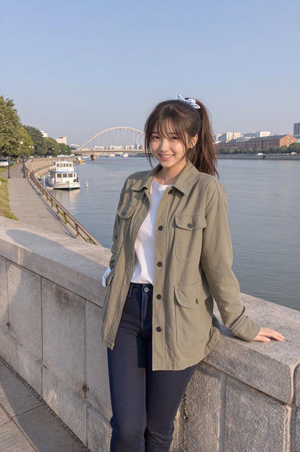 강변 돌담에 기대 웃고 있는 Riho. 올리브색 재킷에 청바지 차림이고, 뒤로 넓은 강과 아치형 다리, 작은 유람선이 보인다